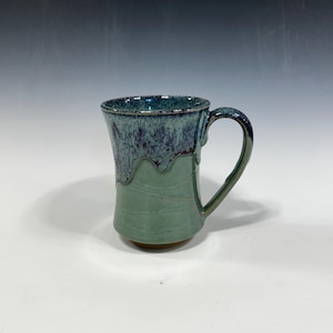 May include: A handmade ceramic mug with a unique design. The mug features a dark teal rim and handle, transitioning to a speckled teal and purple upper section. The lower half is a muted green with horizontal lines. The mug sits on a small, round base.