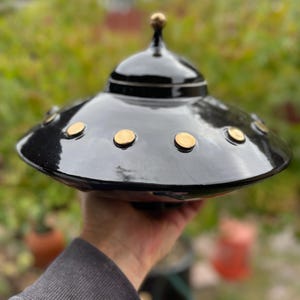 May include: A black ceramic UFO-shaped decorative piece with gold-colored accents. The top has a small gold knob, and the base has several gold circles. The piece is glossy and appears to be handmade.