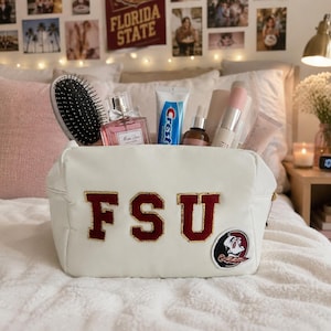 May include: A white cosmetic bag with the letters "FSU" in maroon and gold. The bag is filled with beauty products, including a hairbrush, perfume, and toothpaste. The bag is on a bed with a white comforter.