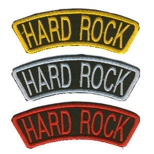 Könnte beinhalten: Drei gestickte Aufnäher mit dem Text "HARD ROCK". Der obere Aufnäher hat einen gelben Rand und Schriftzug, der mittlere einen silbernen Rand und Schriftzug, und der untere einen roten Rand und Schriftzug. Alle Aufnäher sind auf schwarzem Hintergrund.