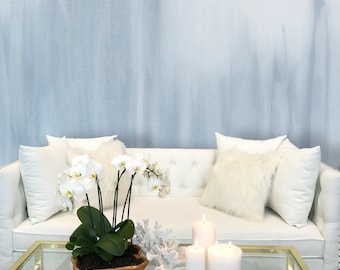 May include: A white couch with white pillows and a white orchid plant in a wooden pot on a glass coffee table with three white candles. The background is a light blue wall.