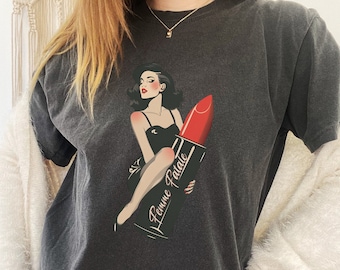 Può includere: Una maglietta grigio scuro con una grafica di una donna in un vestito nero seduta su un grande rossetto rosso. Il rossetto ha le parole "Femme Fatale" scritte su di esso.