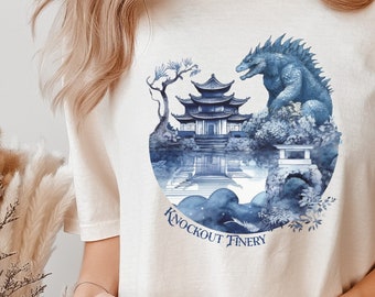 May include: A cream colored t-shirt with a blue watercolor design of a Godzilla-like monster standing in front of a Japanese temple. The text "Knockout Finery" is printed below the design.