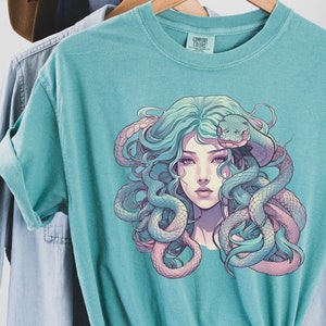 Puede incluir: Una camiseta azul turquesa con un estampado gráfico de una mujer con serpientes en el pelo. La mujer tiene el pelo largo y verde y lleva una serpiente rosa y morada alrededor de la cabeza. Las serpientes son verdes y rosas.
