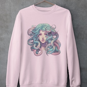 Puede incluir: Una sudadera rosa con un estampado gráfico de una mujer con serpientes en el pelo. La mujer tiene el pelo azul y una serpiente azul claro envuelta alrededor de su cabeza.