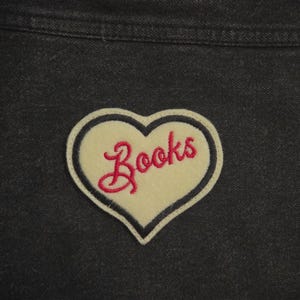 May include: A heart-shaped patch with the word "Books" embroidered in pink cursive. The patch has a cream-colored center, a black border, and is set against a dark denim background.
