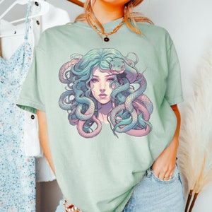 Pode incluir: Uma camiseta verde claro com uma estampa gr&aacute;fica de uma mulher com cobras no cabelo. A mulher tem cabelo longo e verde e uma cobra azul claro enrolada em torno de sua cabe&ccedil;a. A cobra tem a barriga rosa.
