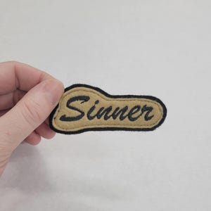 May include: A tan felt patch with black stitching and the word "Sinner" embroidered in black.