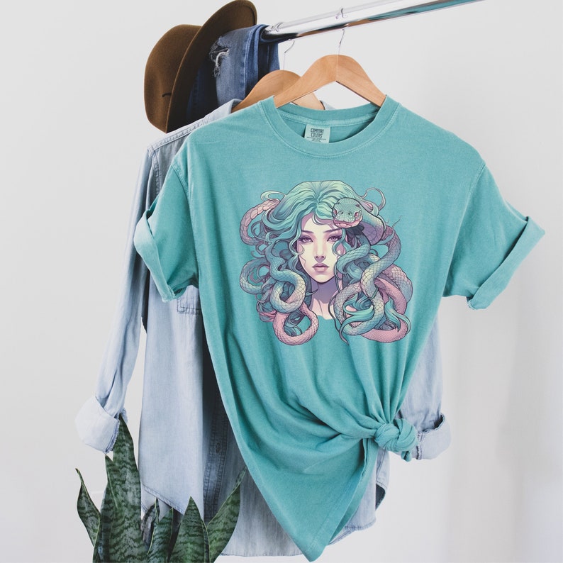Pode incluir: Uma camiseta azul turquesa com uma estampa gr&aacute;fica de uma mulher com cobras no cabelo. A mulher tem cabelo comprido e verde e usa uma cobra rosa e roxa em volta da cabe&ccedil;a. As cobras s&atilde;o verdes e rosas.
