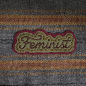 Op de afbeelding: Een stoffen patch met het woord "Feminist" in groen schrift, omlijnd in goud en rood. De patch is geplaatst tegen een grijze stoffen achtergrond met horizontale strepen in oranje en bruin.