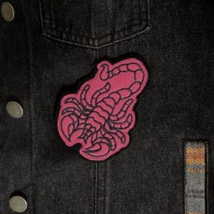 May include: A pink scorpion patch with black outline is sewn onto a black denim jacket. The patch is a stylized illustration of a scorpion. The jacket has silver buttons and a pocket.