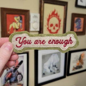 May include: A green felt patch with white stitching in the shape of a bone. The patch has the words "You are enough" embroidered in red thread.