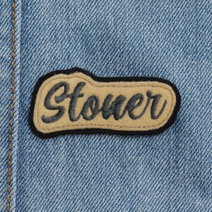Puede incluir: Primer plano de una chaqueta vaquera azul claro con un parche cosido. El parche es de color beige con texto bordado en negro que dice "Stoner" en una fuente estilizada, delineado en negro.
