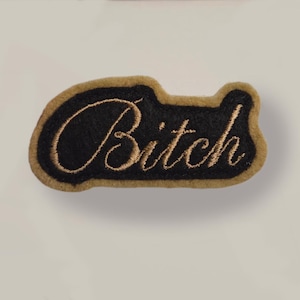 May include: A black patch with the word "Bitch" embroidered in gold script. The patch has a gold border and a unique shape, likely for clothing or accessories.