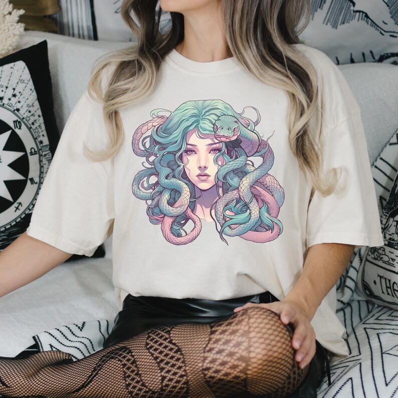 Pode incluir: Uma camiseta branca com uma estampa gr&aacute;fica de uma mulher com cobras no cabelo. A mulher tem cabelo azul e as cobras s&atilde;o verdes e rosas.