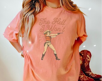May include: A coral pink t-shirt with a vintage-style illustration of a woman in a cowboy hat holding a gun. The text "The Hell I Won't" is printed in brown on the shirt.