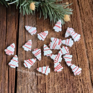 May include: A collection of small, white Christmas tree-shaped embellishments with red stripes and green polka dots. The embellishments are scattered on a wooden surface, with a sprig of evergreen and a decorative ball in the background. The text "DesignsByJNP.com" is visible.