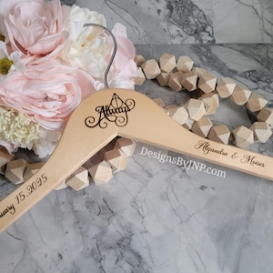 May include: A wooden hanger with the word "Always" and a decorative design, along with the names "Alejandra & Moises" and the date "February 15, 2025" engraved on it. The hanger is light-colored and has a hook. A bouquet of pink flowers and wooden beads are in the background.