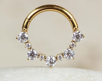 May include: Gold body jewelry with a circular design and clear gemstones. The jewelry is a ring with a row of gemstones around the top.