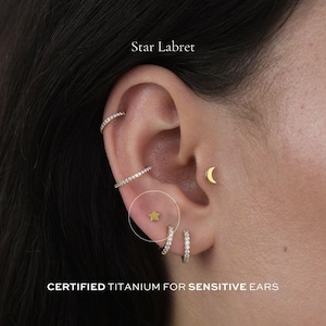 May include: An ear adorned with gold earrings, including a star labret. The image features the text "Star Labret" and "CERTIFIED TITANIUM FOR SENSITIVE EARS". The earrings are designed for sensitive ears.