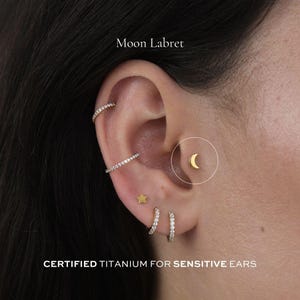 Può includere: Primo piano di un orecchio con diversi orecchini, tra cui un labret a forma di luna crescente dorata. Altri orecchini includono un orecchino a forma di stella dorata, due piccoli orecchini a cerchio e due cerchietti tempestati di diamanti. Il testo recita "Moon Labret" e "CERTIFIED TITANIUM FOR SENSITIVE EARS."