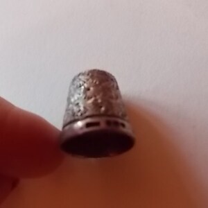Puede incluir: Un dedal de plata con un dise&ntilde;o floral intrincado. El dedal tiene una parte superior lisa y redondeada y una base ligeramente acampanada.