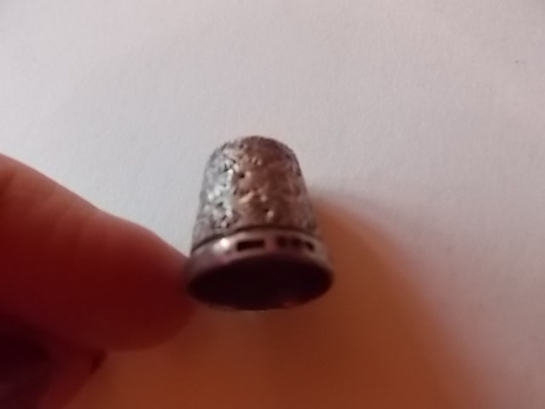 Puede incluir: Un dedal de plata con un dise&ntilde;o floral intrincado. El dedal tiene una parte superior lisa y redondeada y una base ligeramente acampanada.