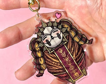 Peut inclure: Un porte-clés en acrylique transparent avec un personnage de dessin animé vêtu de marron et d'or, avec un visage blanc et des yeux noirs. Le porte-clés a un fermoir doré en forme de cœur et une bannière avec du texte.