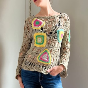 May include: A beige crochet sweater with colorful geometric patches. The sweater has a loose fit and long sleeves. The sweater is made of a lightweight material and has a distressed look.
