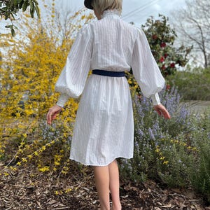 K&ouml;nnte beinhalten: Ein wei&szlig;es Kleid mit einem gestreiften Muster und einem R&uuml;schenkragen. Das Kleid hat lange &Auml;rmel mit Puff&auml;rmeln und einen dunkelblauen G&uuml;rtel. Das Model tr&auml;gt schwarz-wei&szlig;e Schuhe.