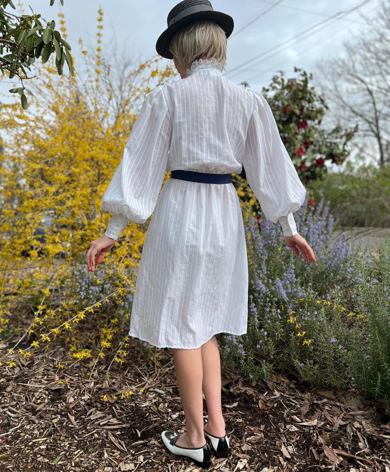 K&ouml;nnte beinhalten: Ein wei&szlig;es Kleid mit einem gestreiften Muster und einem R&uuml;schenkragen. Das Kleid hat lange &Auml;rmel mit Puff&auml;rmeln und einen dunkelblauen G&uuml;rtel. Das Model tr&auml;gt schwarz-wei&szlig;e Schuhe.