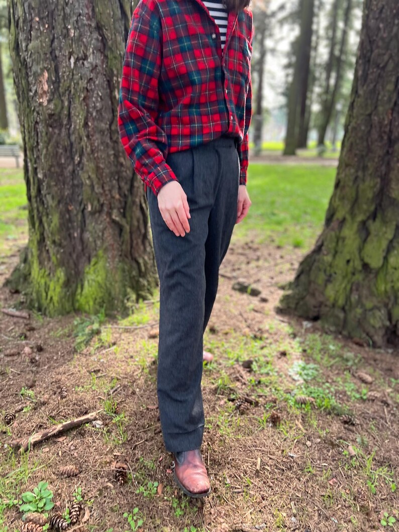 May include: A person wearing a red and green plaid flannel shirt over a black and white striped shirt, paired with dark gray trousers and brown leather boots. The outfit is casual and suitable for outdoor activities.