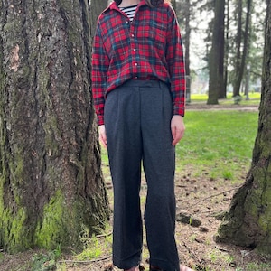 May include: A person wearing a red and green plaid flannel shirt over a black and white striped shirt, paired with dark gray wide-leg pants. The outfit is completed with brown shoes. The setting is outdoors, with trees in the background.