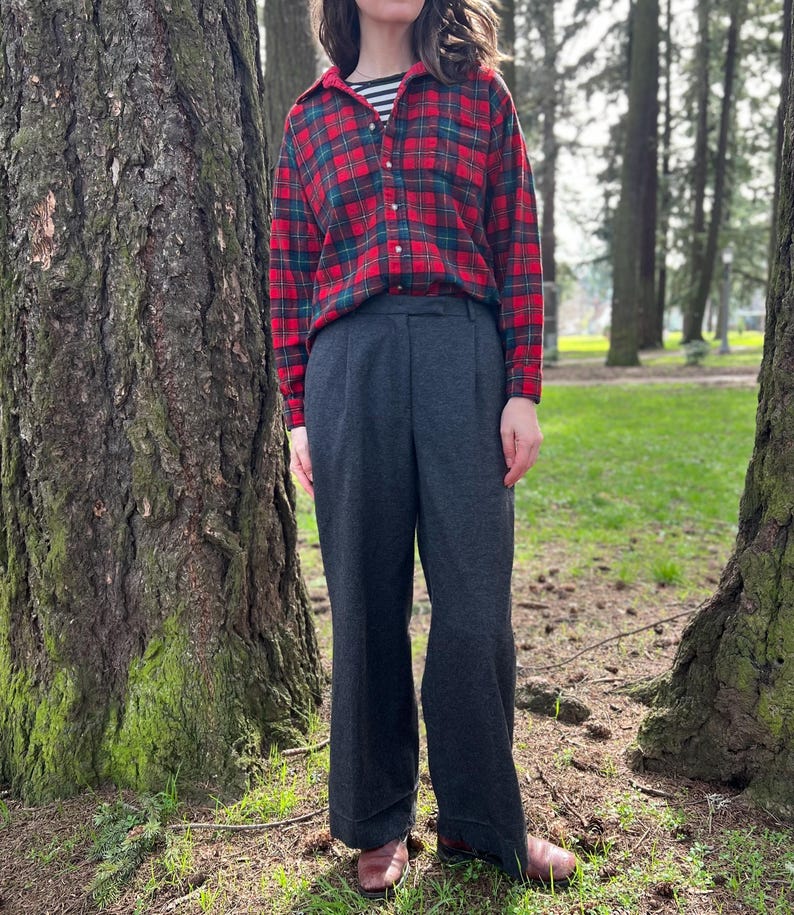 May include: A person wearing a red and green plaid flannel shirt over a black and white striped shirt, paired with dark gray wide-leg pants. The outfit is completed with brown shoes. The setting is outdoors, with trees in the background.