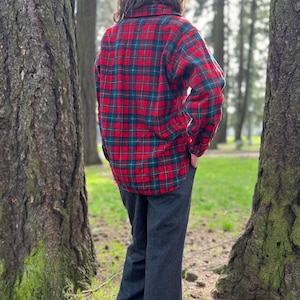 May include: A red, green, and black plaid flannel shirt. The shirt has long sleeves and a button-down front. The person is wearing dark gray pants. The image is taken outdoors, with trees in the background.