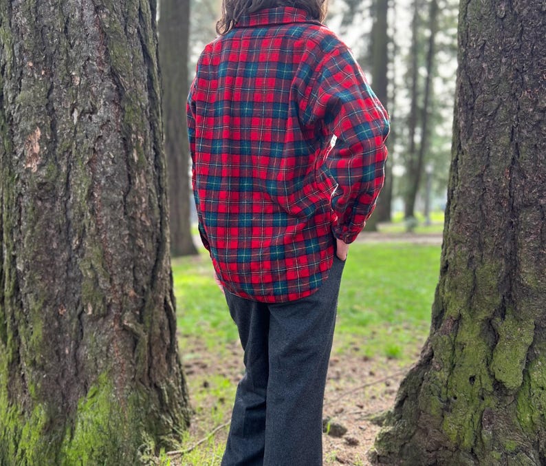 May include: A red, green, and black plaid flannel shirt. The shirt has long sleeves and a button-down front. The person is wearing dark gray pants. The image is taken outdoors, with trees in the background.