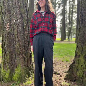 May include: A person wearing a red and green plaid flannel shirt over a black and white striped shirt, dark gray pleated trousers, a black cap, and sunglasses. The person is standing in a park with large trees.