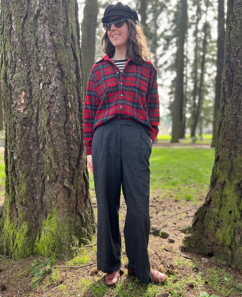May include: A person wearing a red and green plaid flannel shirt over a black and white striped shirt, dark gray pleated trousers, a black cap, and sunglasses. The person is standing in a park with large trees.