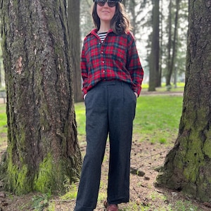 May include: A person wearing a red and black plaid shirt, a black cap, and dark sunglasses. The person is also wearing a striped shirt underneath the plaid shirt and dark gray pants. The person is standing in a park with trees in the background.