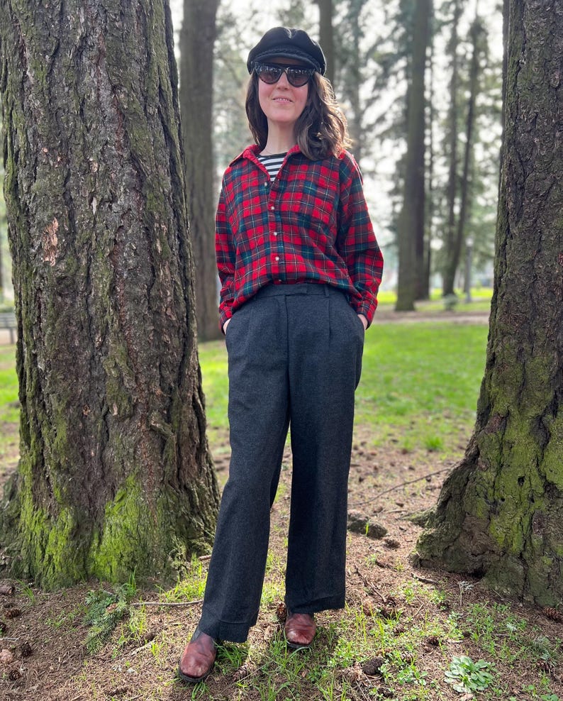 May include: A person wearing a red and black plaid shirt, a black cap, and dark sunglasses. The person is also wearing a striped shirt underneath the plaid shirt and dark gray pants. The person is standing in a park with trees in the background.