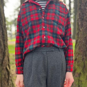 May include: A red and green plaid flannel shirt with a button-down front is paired with gray trousers. The shirt is layered over a black and white striped top. The outfit is set against a blurred outdoor background.