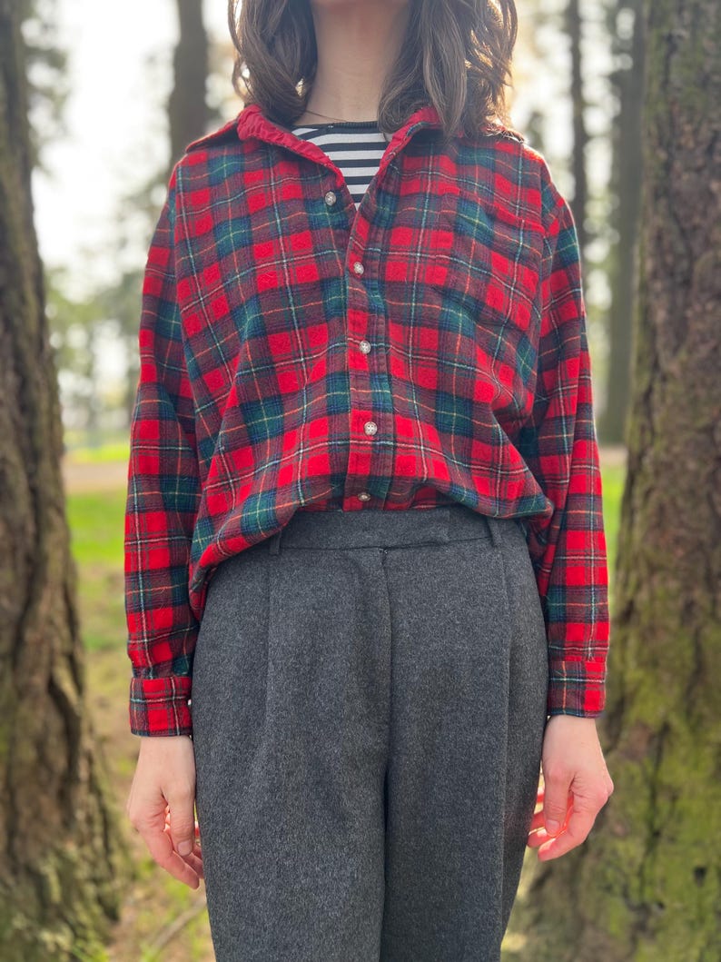 May include: A red and green plaid flannel shirt with a button-down front is paired with gray trousers. The shirt is layered over a black and white striped top. The outfit is set against a blurred outdoor background.