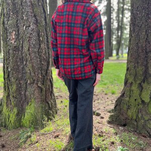 May include: A red and green plaid flannel shirt with long sleeves and dark gray pants. The shirt has a classic lumberjack style, and the pants have a wide leg. The person is standing in a forest setting.