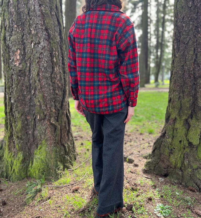 May include: A red and green plaid flannel shirt with long sleeves and dark gray pants. The shirt has a classic lumberjack style, and the pants have a wide leg. The person is standing in a forest setting.