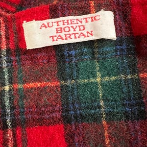 May include: Close-up of a red, green, and blue plaid fabric with a white label that reads "AUTHENTIC BOYD TARTAN" in red letters. The tartan pattern features a mix of red, green, blue, and yellow lines.