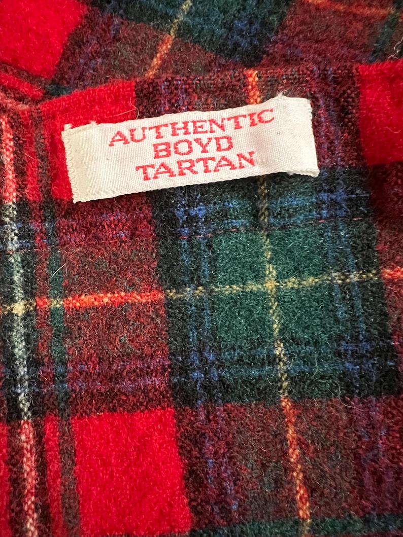 May include: Close-up of a red, green, and blue plaid fabric with a white label that reads "AUTHENTIC BOYD TARTAN" in red letters. The tartan pattern features a mix of red, green, blue, and yellow lines.