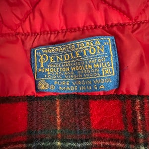 May include: Close-up of a red quilted fabric and a blue label. The label reads "WARRANTED TO BE A PENDLETON" with additional text about the Pendleton Woolen Mills and 100% virgin wool. The bottom portion shows a red and black plaid pattern.