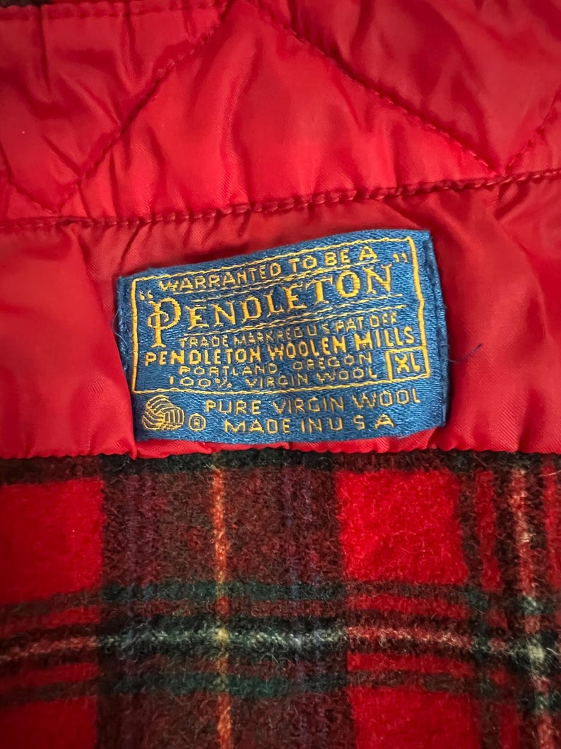 May include: Close-up of a red quilted fabric and a blue label. The label reads "WARRANTED TO BE A PENDLETON" with additional text about the Pendleton Woolen Mills and 100% virgin wool. The bottom portion shows a red and black plaid pattern.