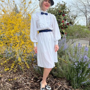 K&ouml;nnte beinhalten: Eine Frau in einem wei&szlig;en Kleid mit R&uuml;schenkragen und einer schwarzen Schleife. Sie tr&auml;gt au&szlig;erdem einen schwarzen Hut und schwarz-wei&szlig;e Slipper. Sie steht vor einem Busch mit gelben Bl&uuml;ten.
