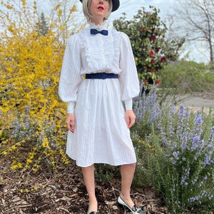 K&ouml;nnte beinhalten: Ein wei&szlig;es Kleid mit R&uuml;schenkragen und langen &Auml;rmeln. Das Kleid hat einen schwarzen G&uuml;rtel und wird mit einem schwarzen Hut und schwarz-wei&szlig;en Schuhen getragen.